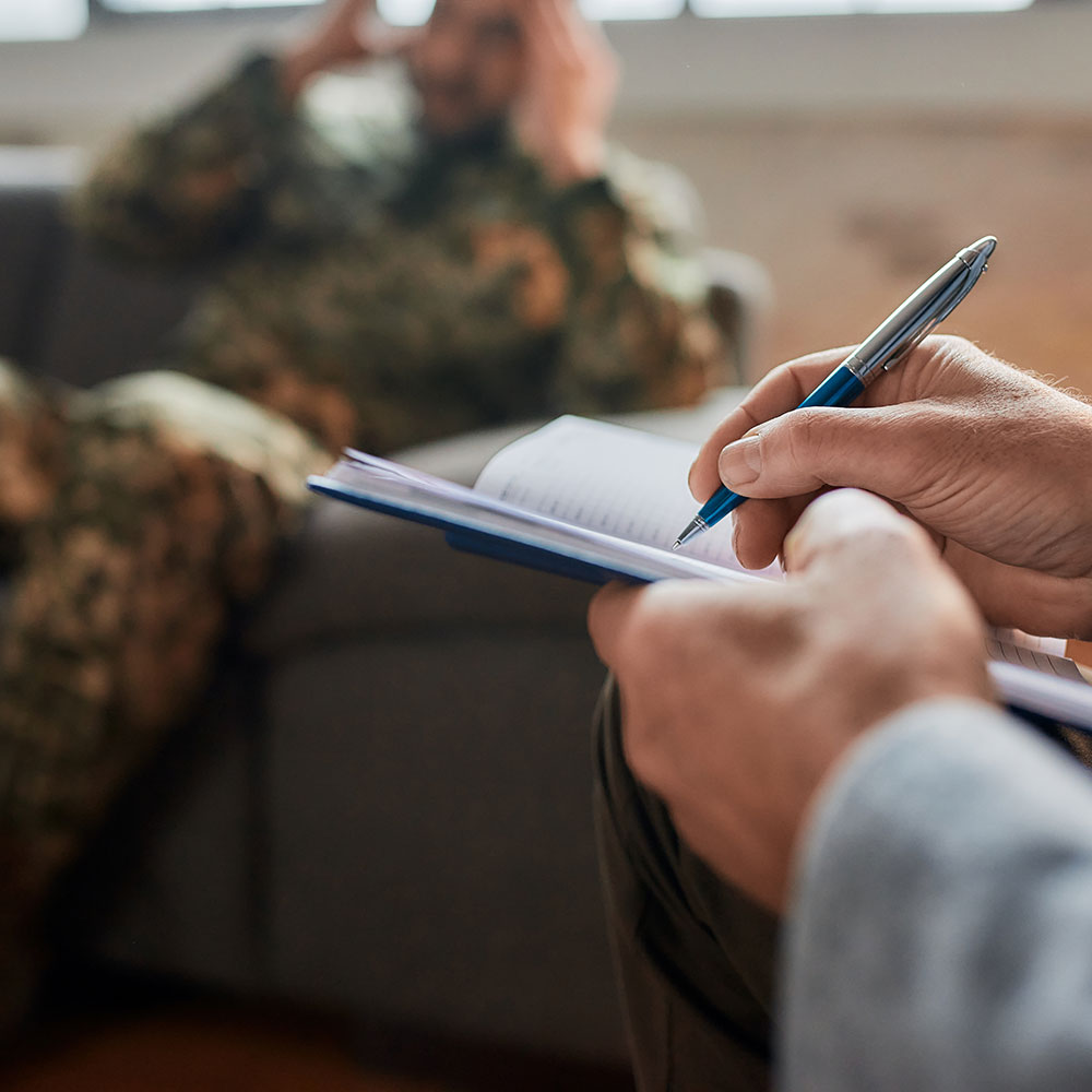 Close up of hands of psychologist making notes, communicating with military man during therapy session. Soldier suffering from depression, psychological trauma. PTSD concept. Focus on hand with pen